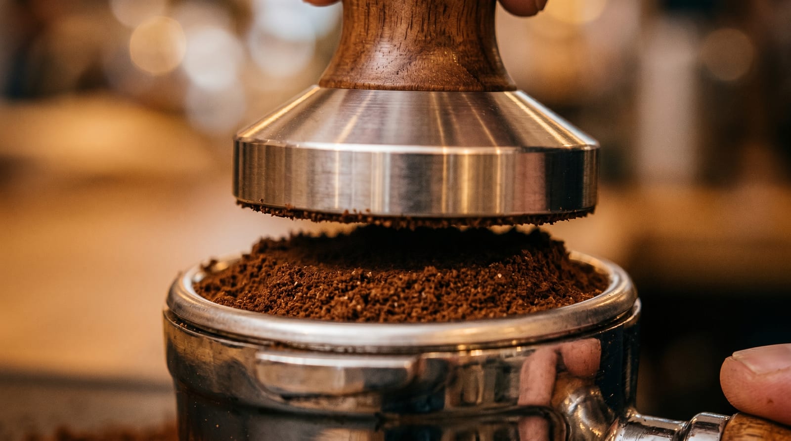 A close-up of a tamper pressing fresh-ground coffee into a stainless-steel portafilter